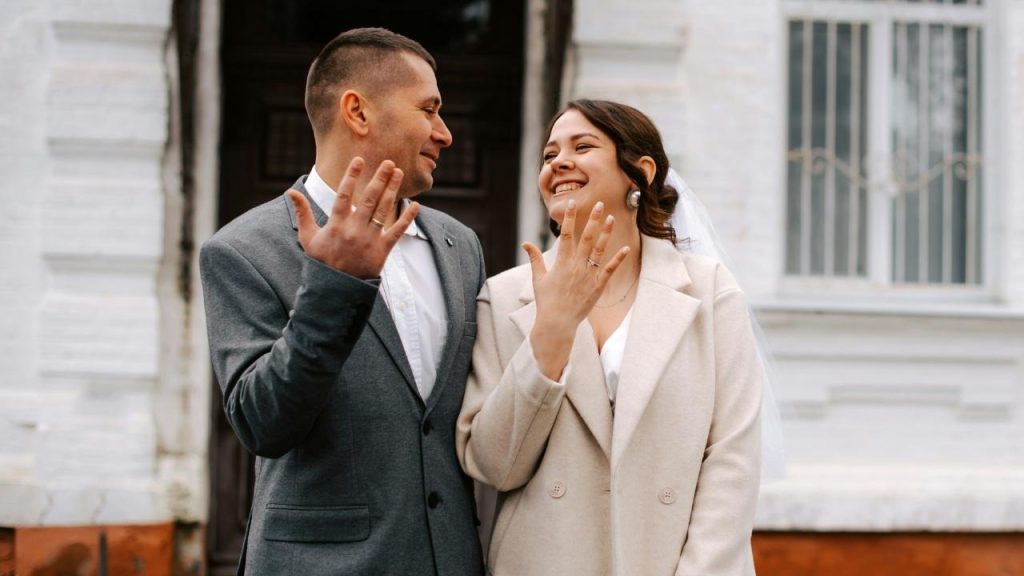 A smiling man and woman stand outside holding up their hands to show rings.