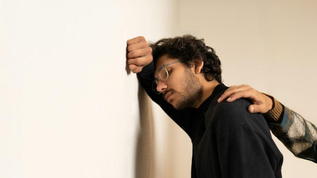 A man leans his head against a white wall while a hand rests on his shoulder.