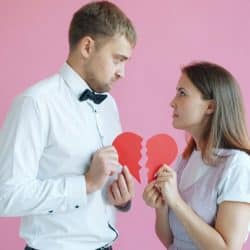 A man and a woman facing each other while holding two halves of a broken red heart against a pink background