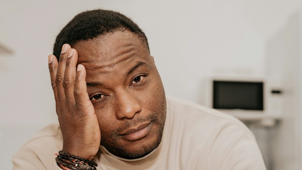 A man resting his head on his hand with a tired expression, wearing a beige shirt and bracelets.