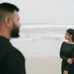 A man and a woman wearing black dresses are standing on a beach separately.