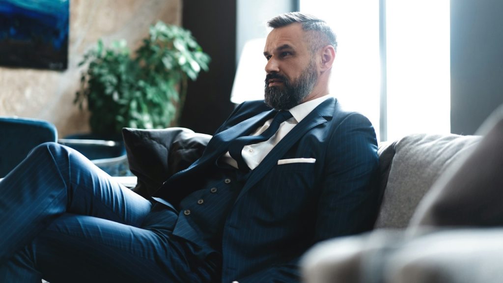 A bearded man in a suit is sitting on a couch.
