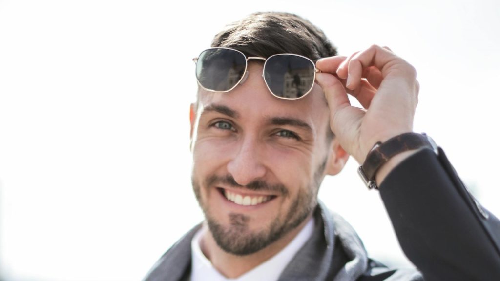 A man in a black jacket, wearing black sunglasses, smiling.