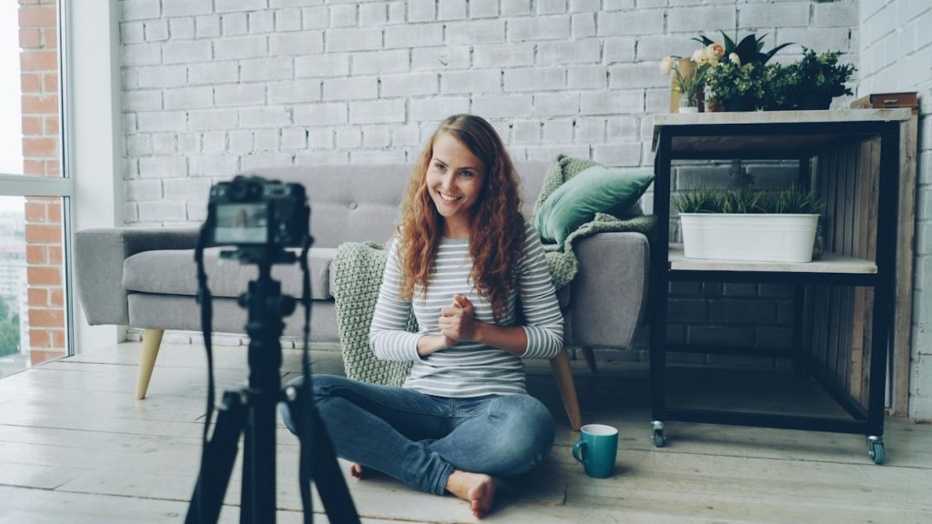 A smiling woman with long curly hair sits cross-legged on the floor of a bright living room, facing a camera on a tripod as if recording a video.