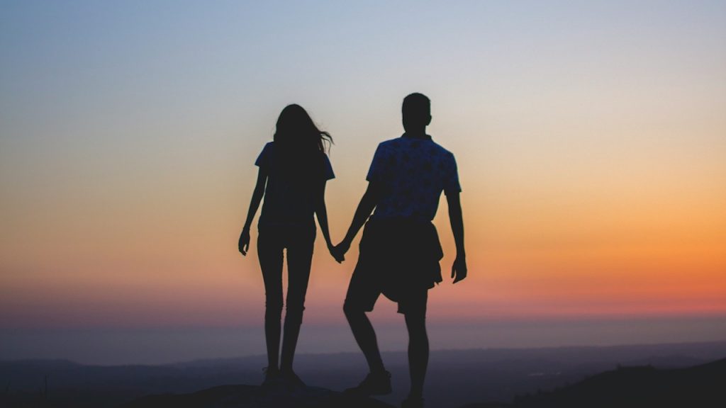 A silhouette of a couple holding hands and standing on a cliff looking at the sunset.