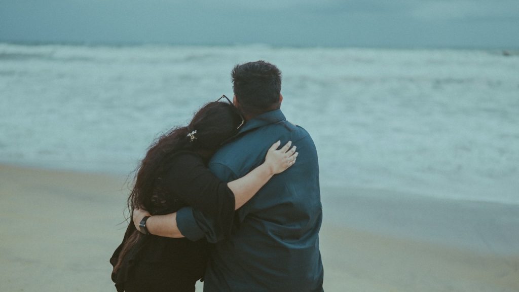 A rearview shot of a couple standing on a sandy beach, looking out at the ocean waves under an overcast sky.