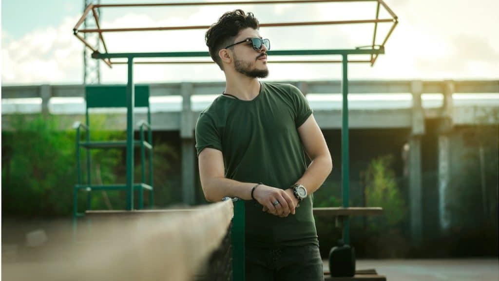 A man in a green crew-neck t-shirt and black pants wearing black sunglasses.