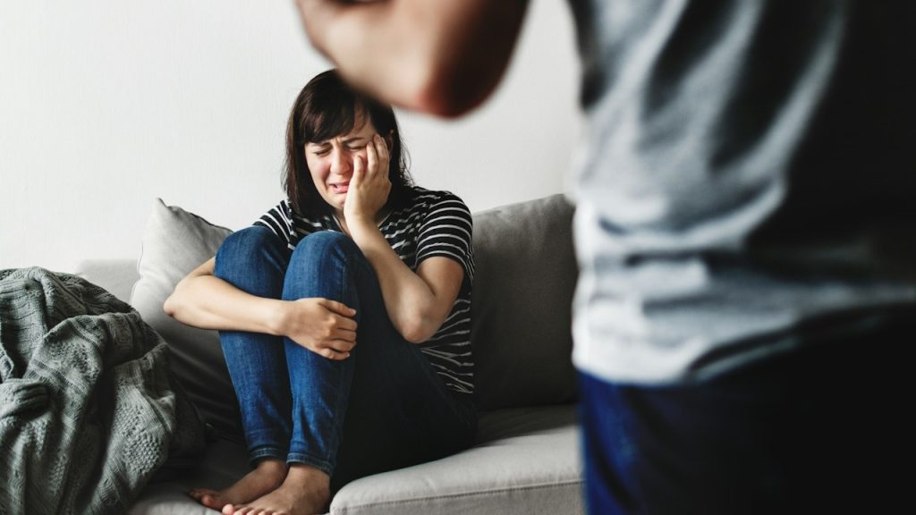 A woman crying while sitting at a couch while her husband is standing in front of her.