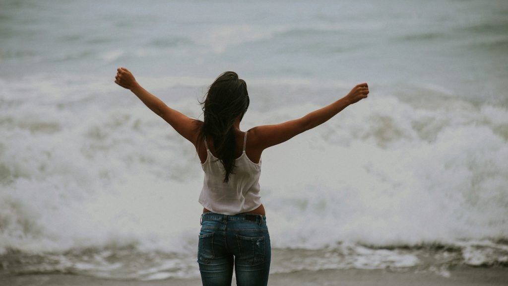 Rear view of a woman standing with arms outstretched facing the crashing ocean waves.
