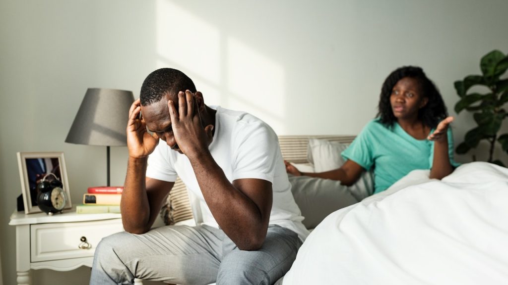 A distressed man sits on the edge of a bed with his head in his hands while a woman gestures argumentatively behind him.