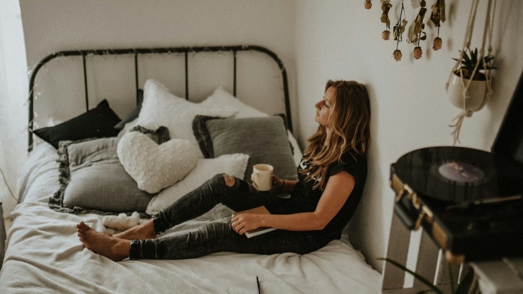 A woman in a black t-shirt and jeans sits on a bed, leaning against the wall with a mug and notebook.