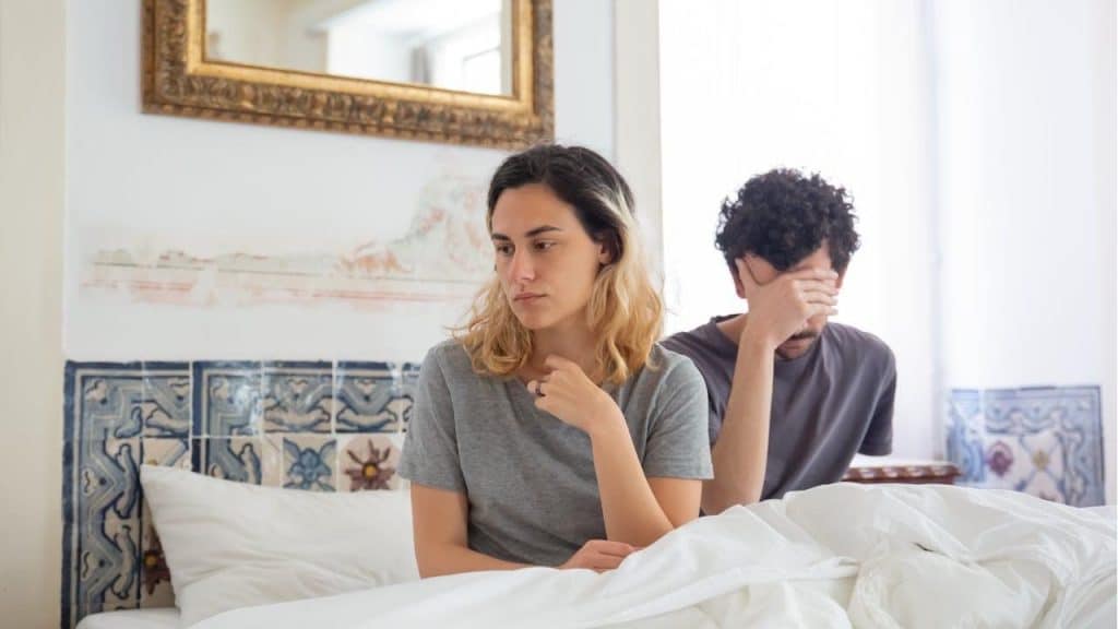 A couple sits  on a bed after an argument; the woman looks sad while the man holds his hand over his face.