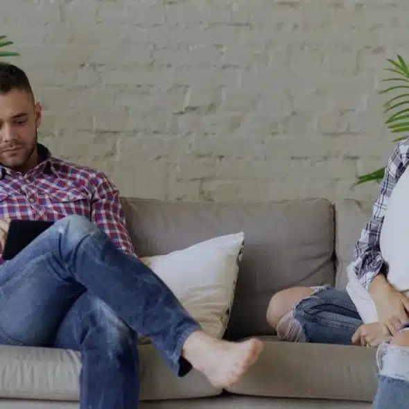 A man is using a tablet while a woman sits on the same couch looking sad, with both ignoring each other.