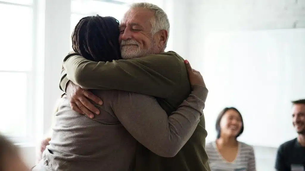 An older man hugging his wife while some people smile in the background.