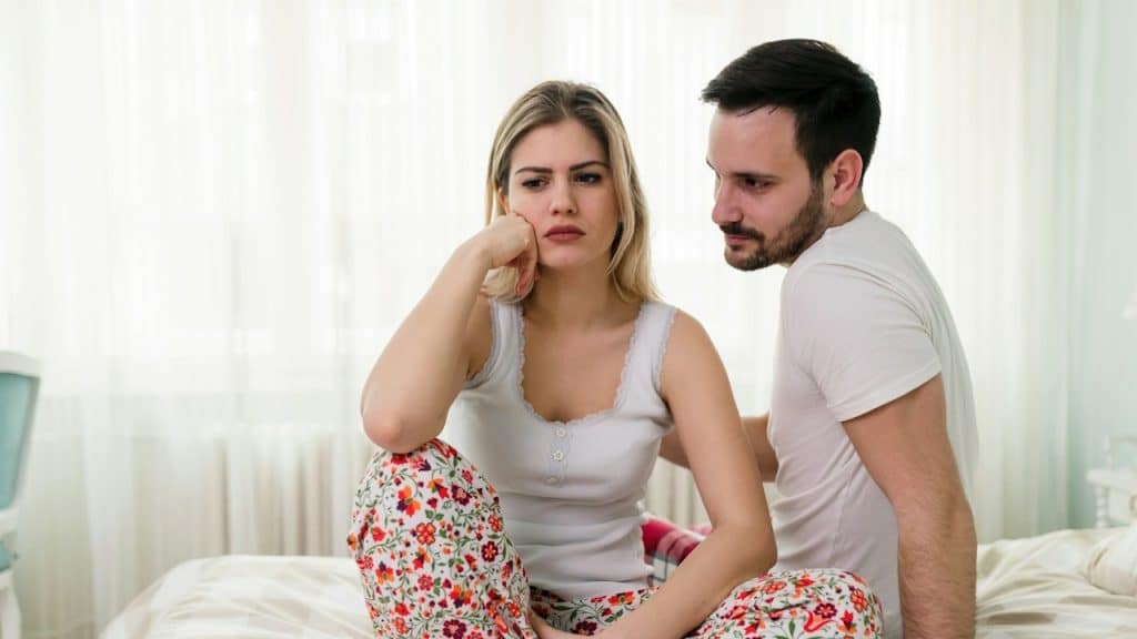 A man looking at a woman appears sad, while they are sitting on their bed.