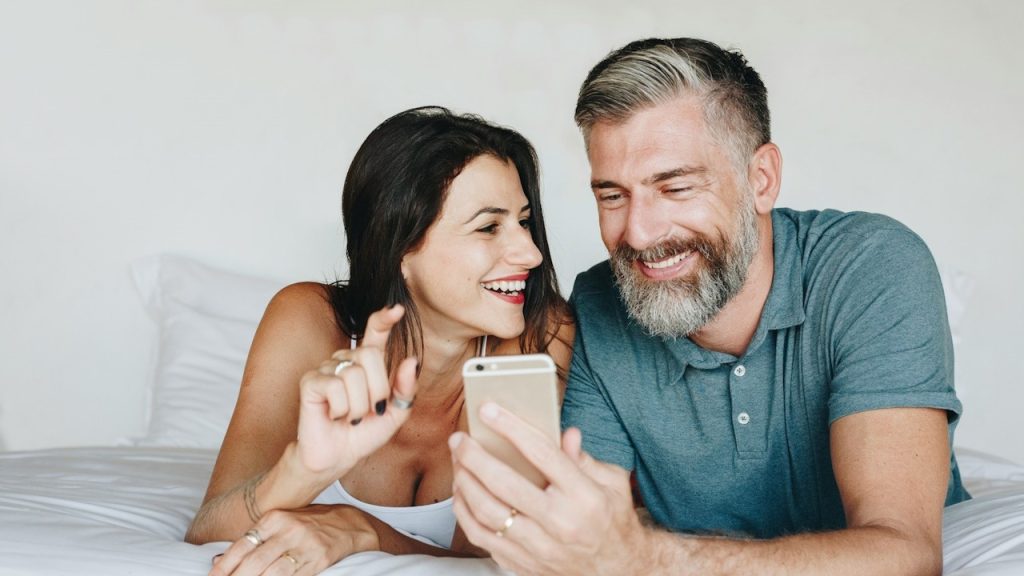 A bearded man with greying beard smiles as he shows something to a woman who looks at him while they lie on a bed.