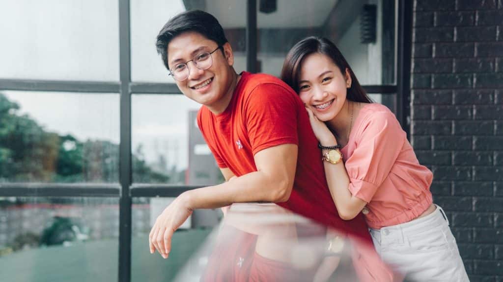 A woman with braces leaning on the back of man wearing a red T-shirt, both smiling on camera.