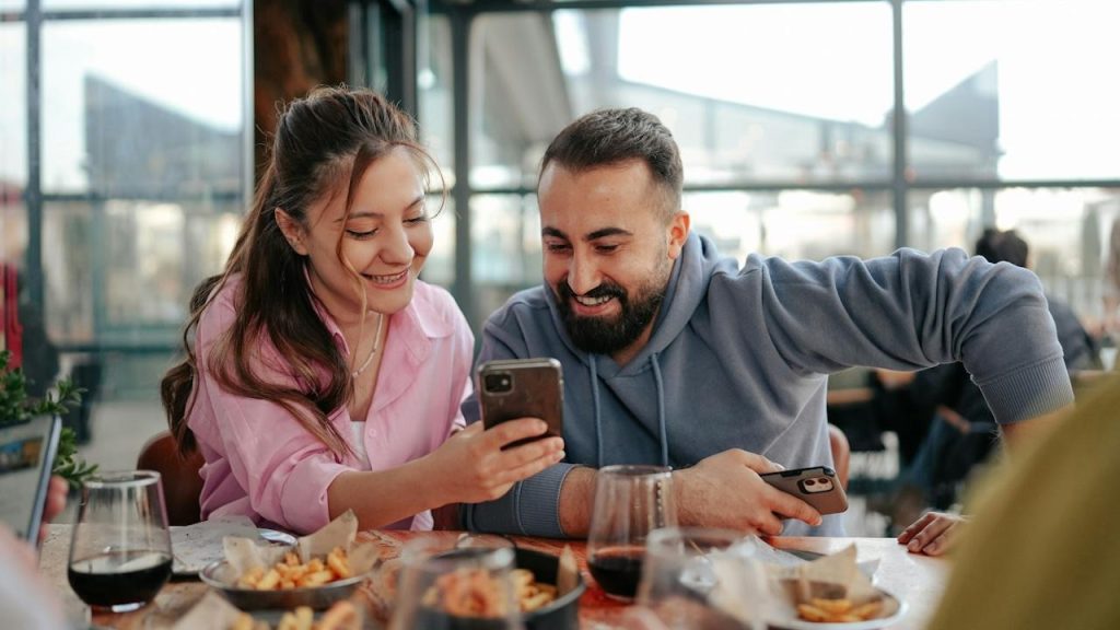 A woman is showing something to her husband on her phone while they are having a meal together.