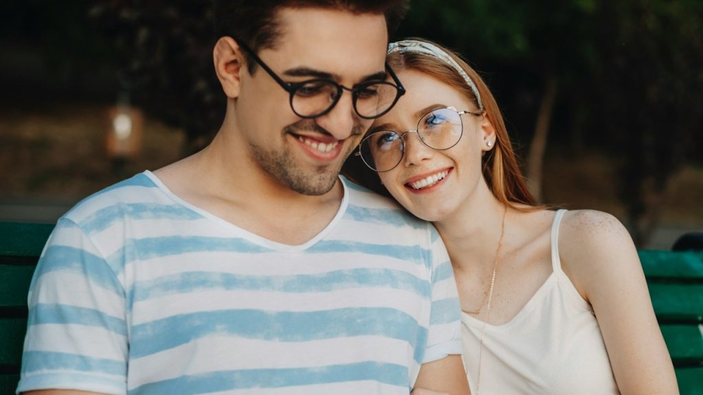 A beautiful couple embracing while sitting on a bench outdoors while a woman with red hair and freckles is looking at him, smiling and leaning her head.