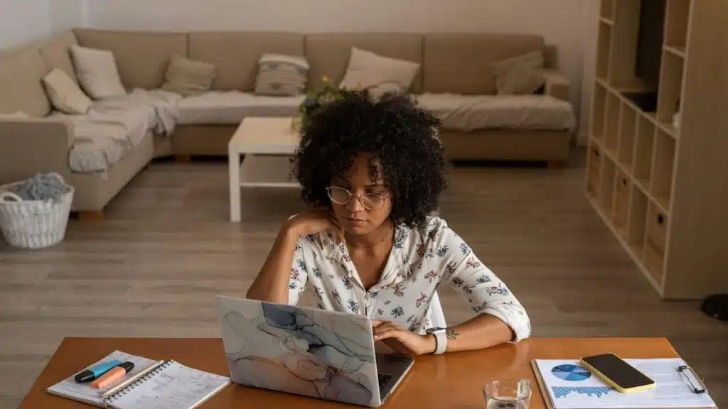 A woman wearing glasses and working on her laptop in her living room.
