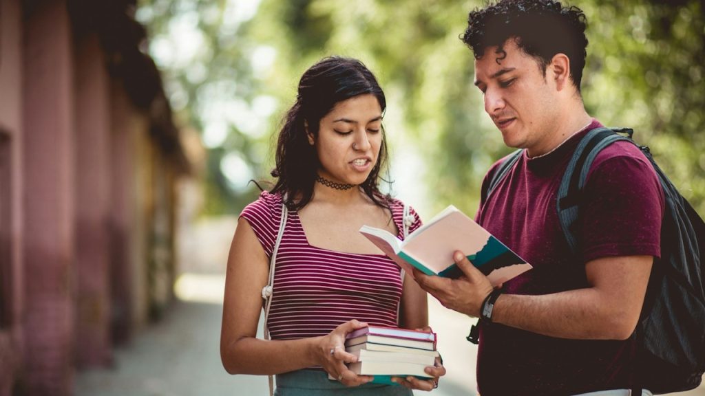 A woman holding some books and looking at a book being held by another man as they stand outside.