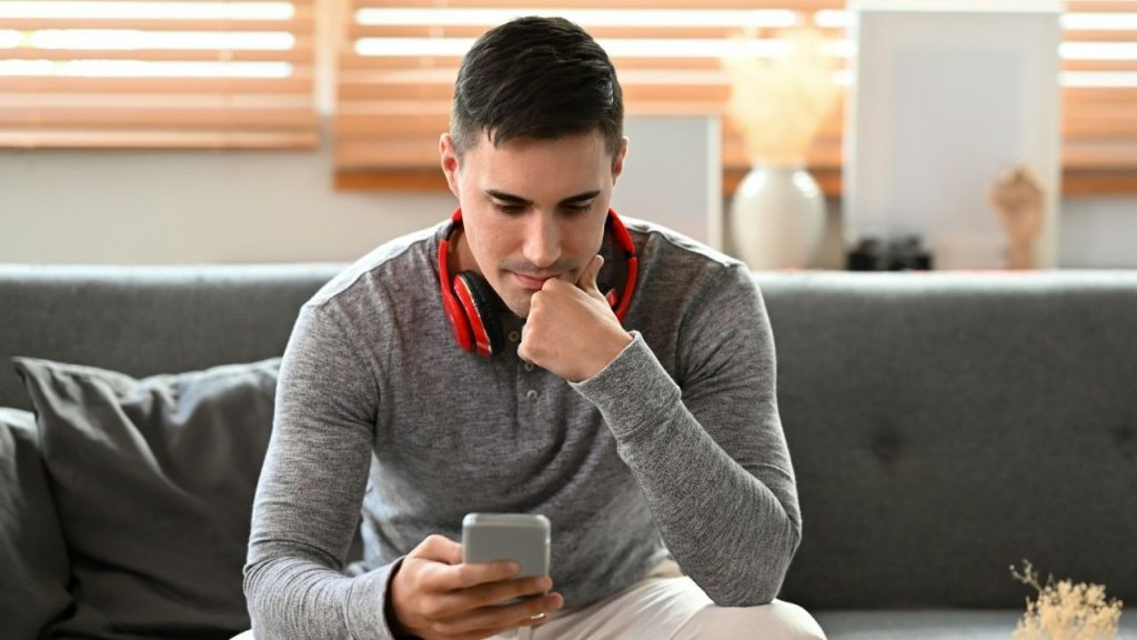 A man with red headphones around his neck looks down at a handheld smartphone screen.