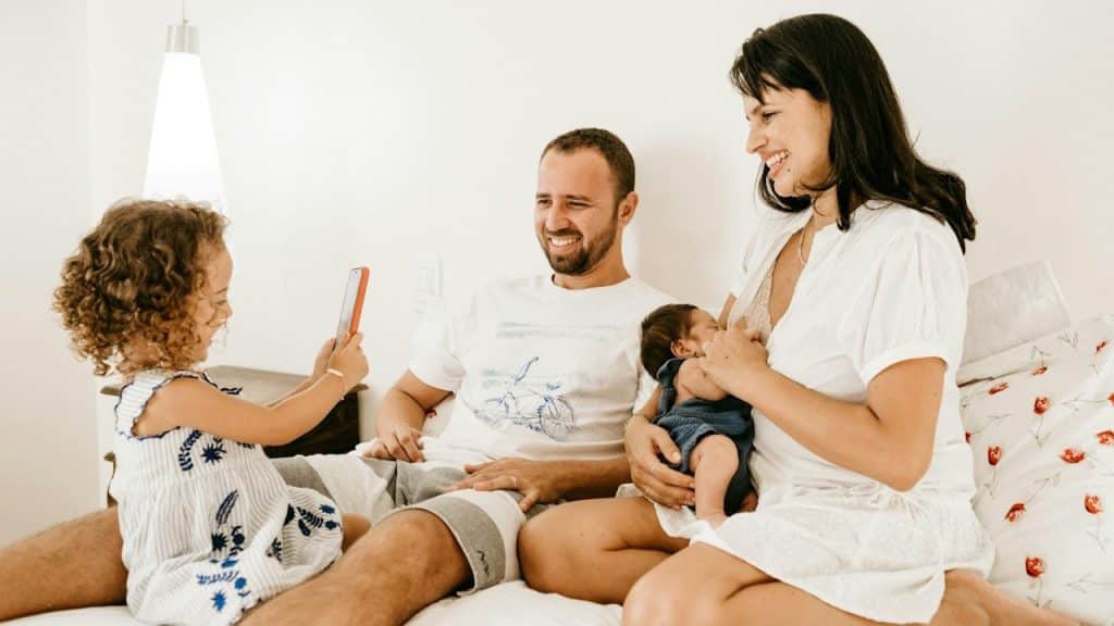A man in a white crew-neck t-shirt sitting beside a woman in a white crew-neck t-shirt is nursing her child and their little daughter is taking their picture.