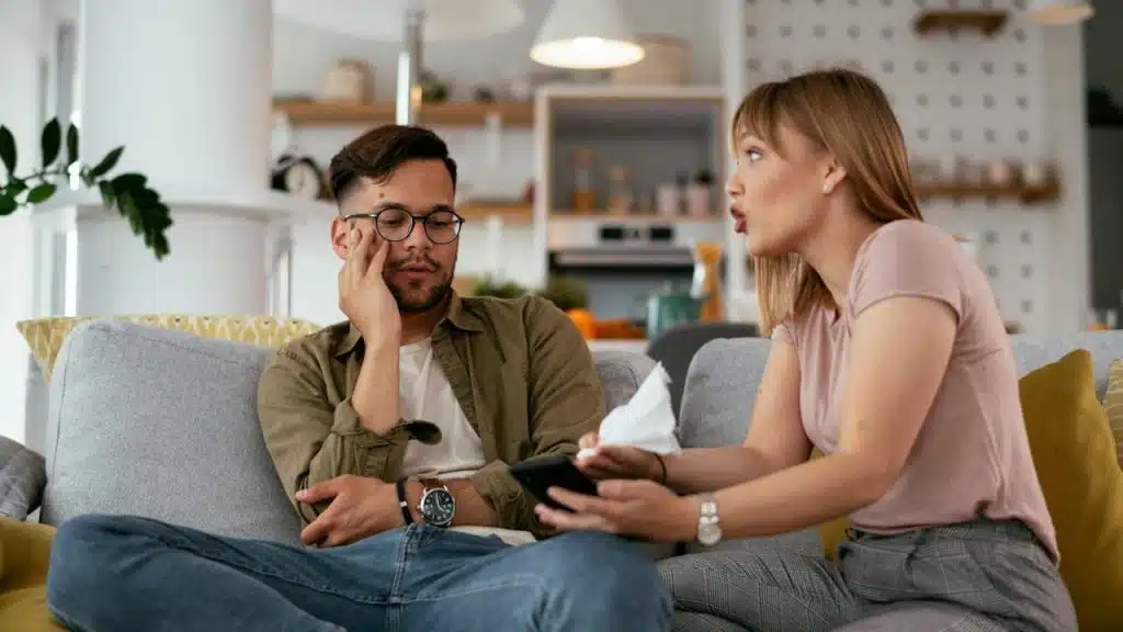 A woman is speaking emphatically to a man who looks distressed as they sit on a couch in a living room.
