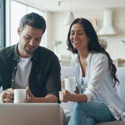 A beautiful young couple drinking coffee and looking at the laptop while sitting on the sofa at home.