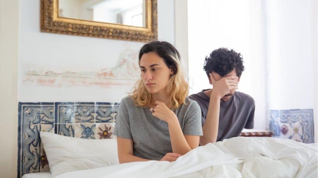 A couple sits on a bed after an argument; the woman looks sad while the man holds his hand over his face.
