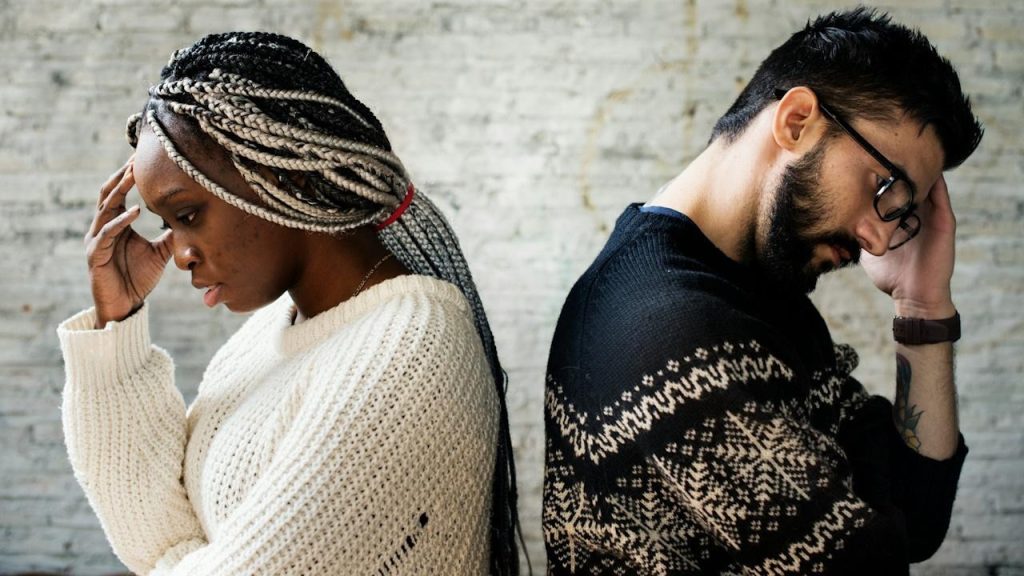 A couple standing with the backs to each other and holding their hands up to their foreheads.