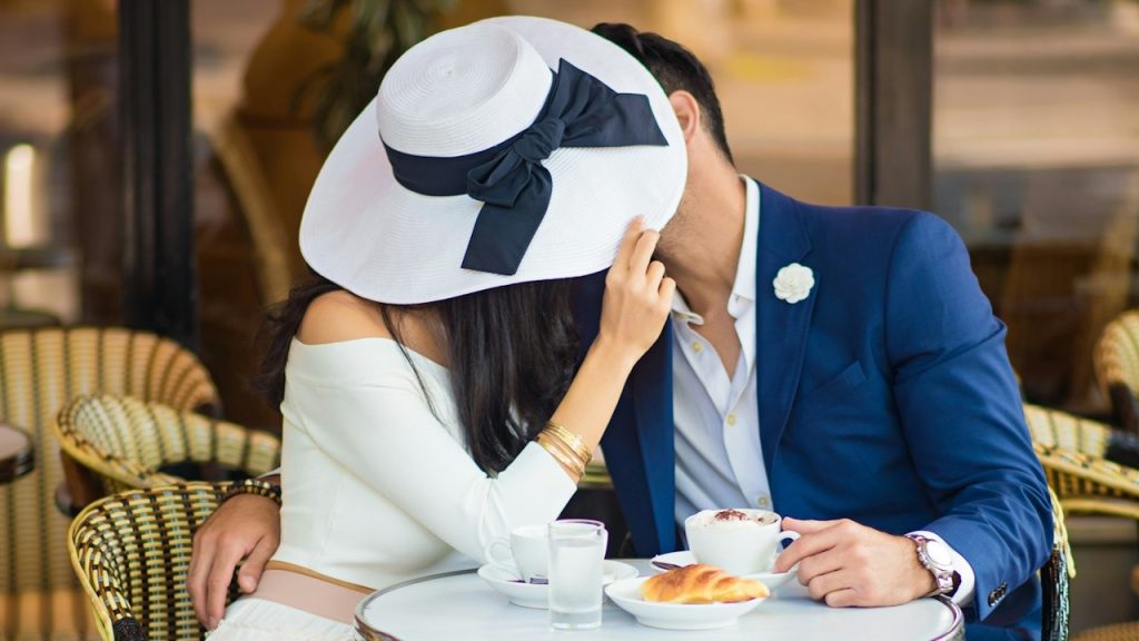 Woman in a white long hat kissing her boyfriend while covering her face with the hat.