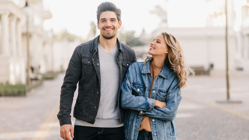 A smiling couple standing on the street while the woman looks at the man.