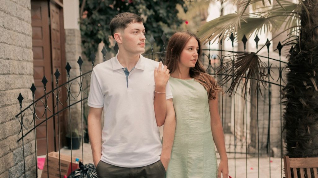A couple standing outdoors with the woman having her ar crossed over his shoulder.
