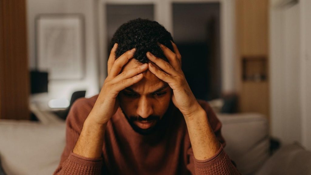 A man sits in a dimly lit room, holding his head in his hands with a stressed expression.