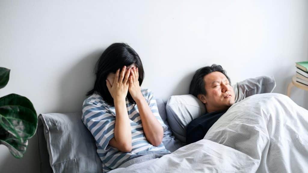 A stressed woman with hands on her face is sitting in bed with a man sleeping beside her.