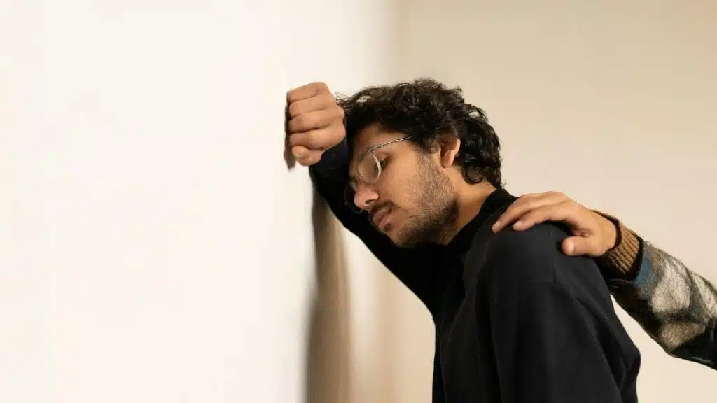 A man leans his head against a white wall while a hand rests on his shoulder.