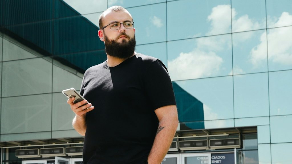A bespectacled and bearded man holding his phone and wearing a black t-shirt standing outside.