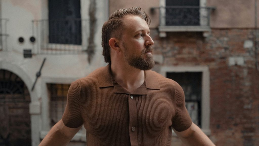 A bearded man wearing a button down shirt standing outside.
