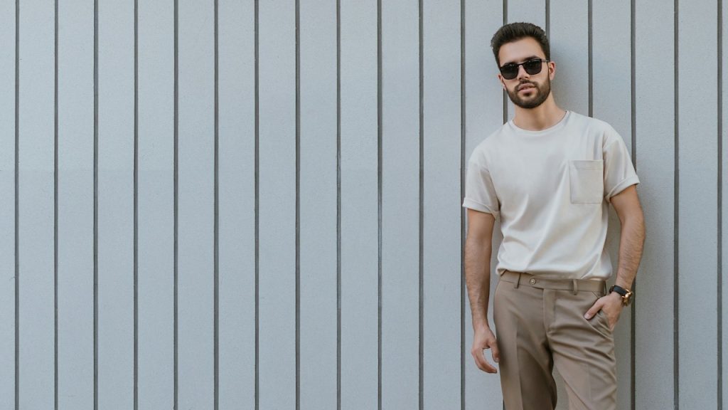 A bearded man wearing sunglasses and chinos leaning on a wall.