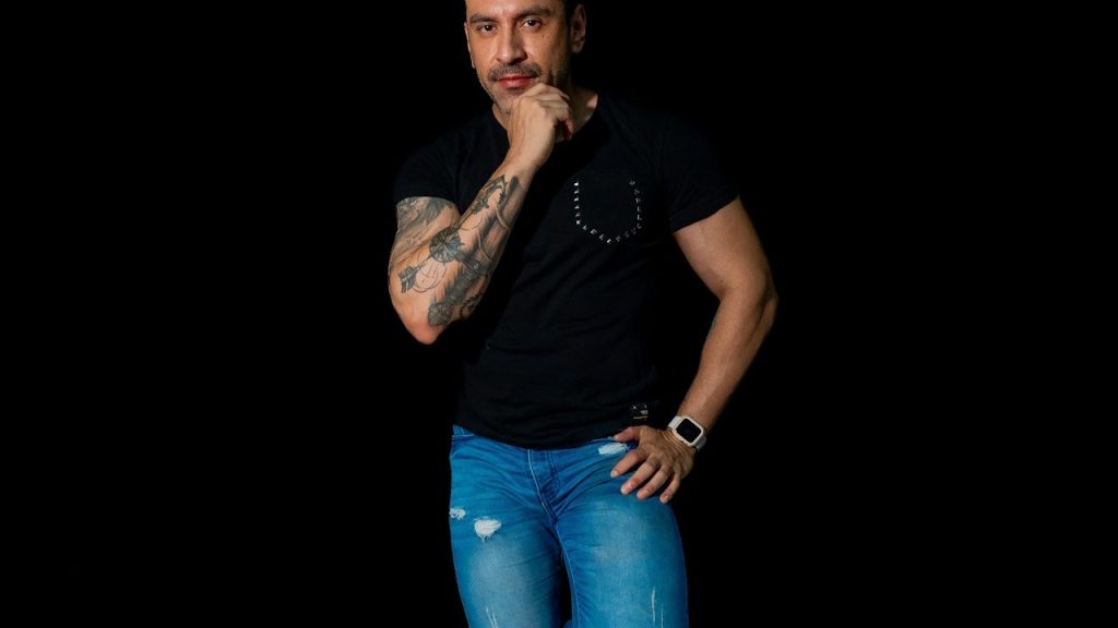 A tattooed man standing with his hand on his chin and wearing a black t-shirt and jeans.