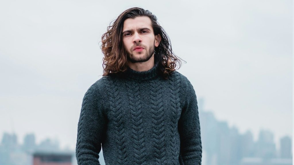 A lightly bearded man with long hair wearing a sweater.