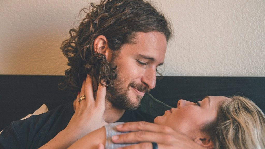 A bearded man is holding a woman who is stroking his beard, and both are looking into each other's eyes.