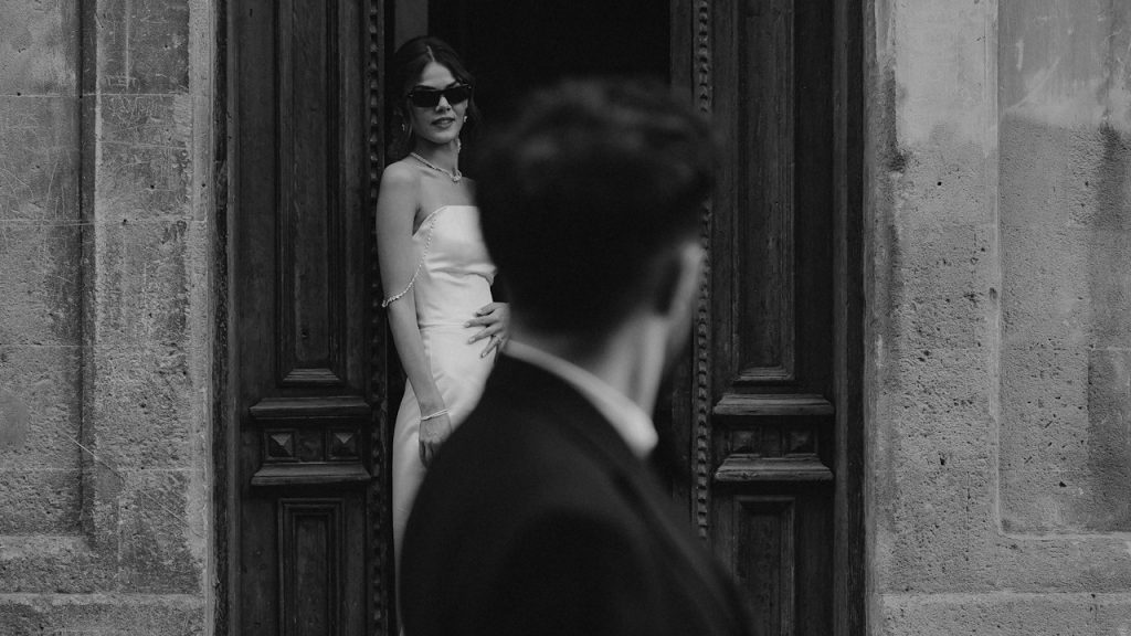 A woman wearing glasses and a white suit looking at a man while standing in a doorway.