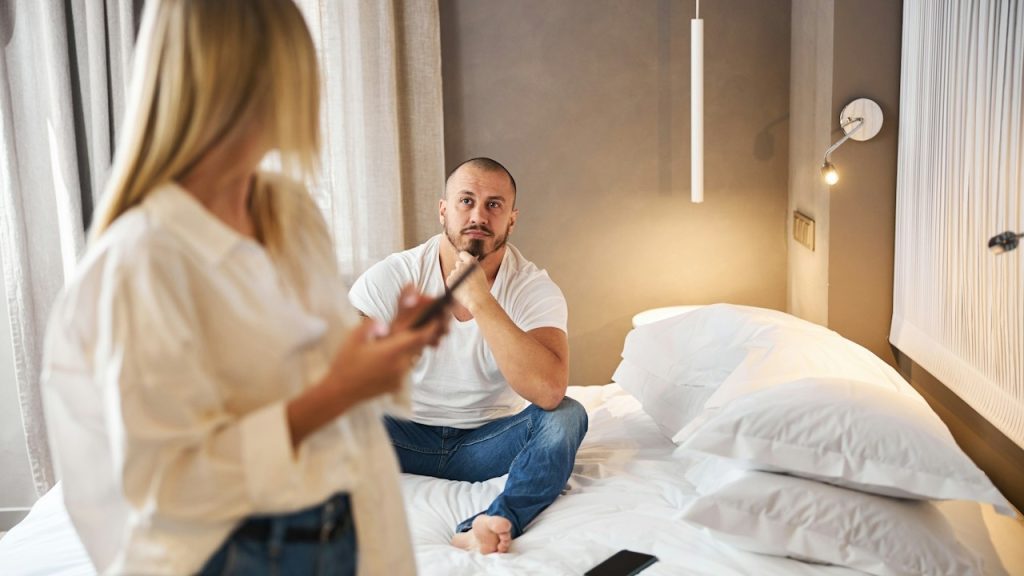 A woman looking back at a bearded man who’s sitting on a bed while using her phone.