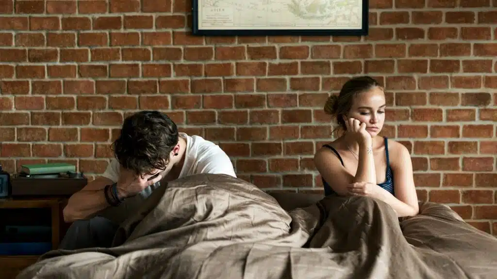 A couple sitting in bed, where the man looks upset and the woman looks surly.