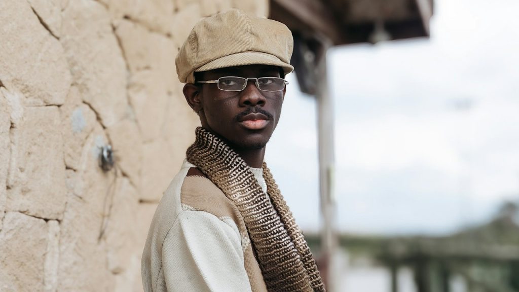 A man in a flat cap and scarf stands near a stone wall.