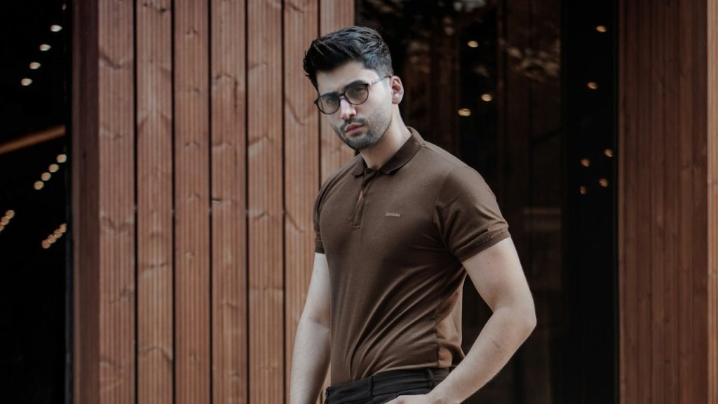 A man in a brown tank top and black pants standing near a brown wooden door.