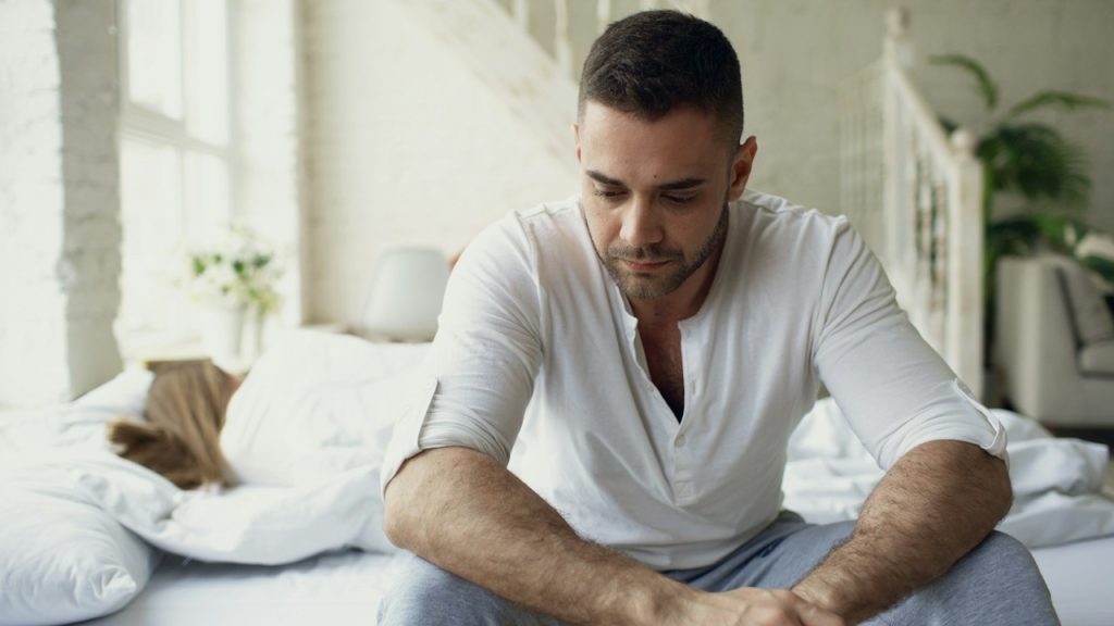 A man sitting on a bed, looking down thoughtfully.