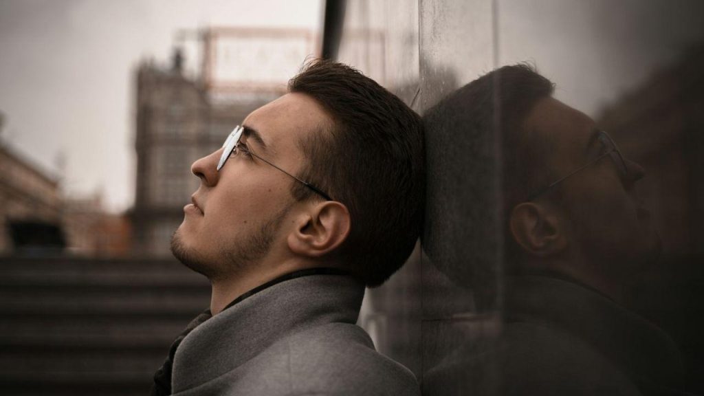 A serious man with glasses and a beard is leaning his head against a stone wall, looking up.